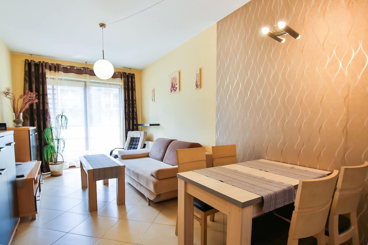 A modern living room with a sofa, dining table, and chairs. There is a kitchen area with cabinets and a countertop.