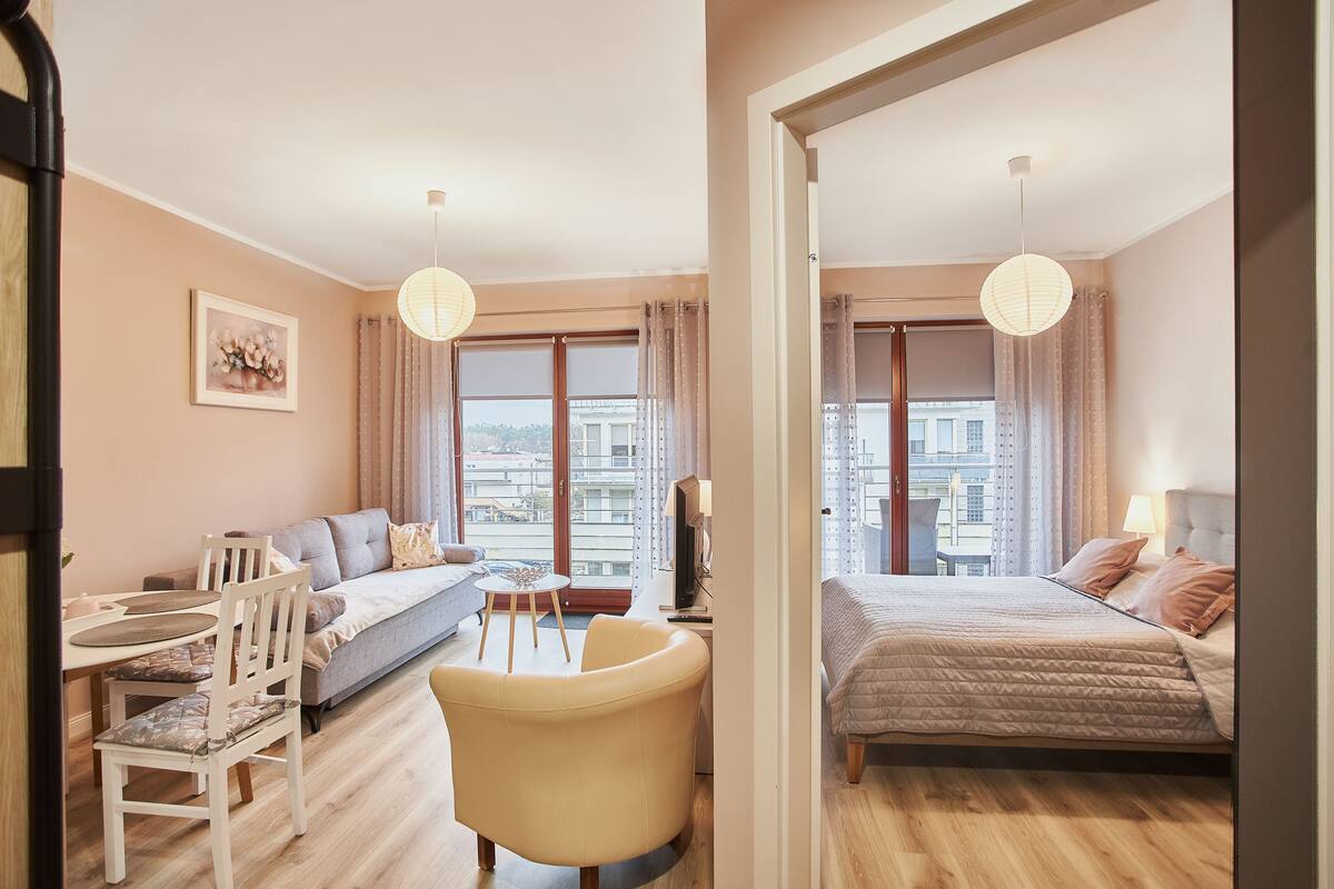 A modern hotel room with a sofa, a dining table, a chair, a bed, and large windows.