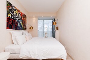 Een slaapkamer met een bed, witte beddengoed, een groot abstract schilderij, een staande lamp en een raam met jaloezieën.