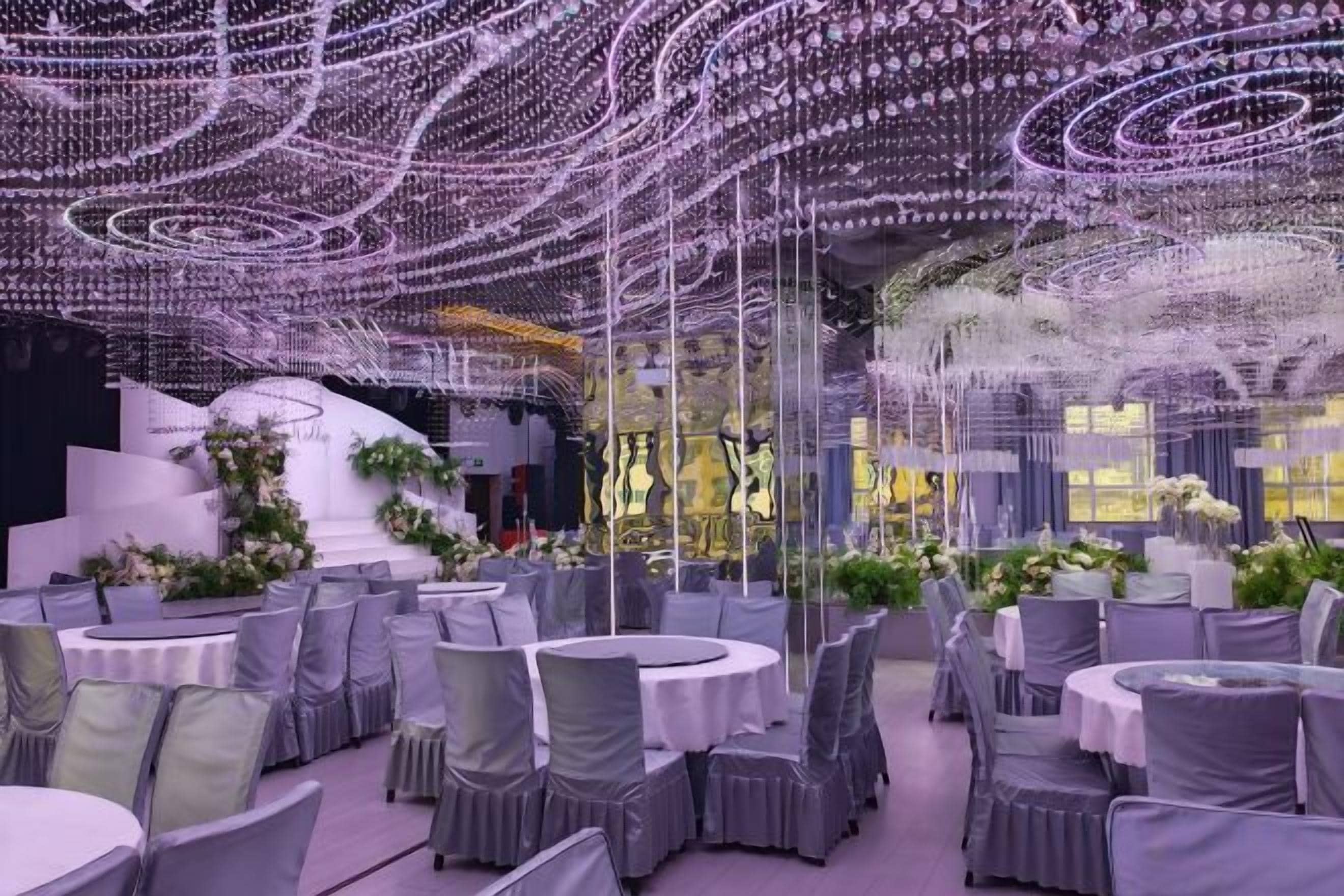A banquet hall with round tables and chairs, a stage with floral arrangements, and a ceiling adorned with string lights.