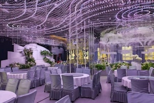 Un salón de banquetes con mesas redondas y sillas, un escenario con arreglos florales y un techo decorado con luces colgantes.