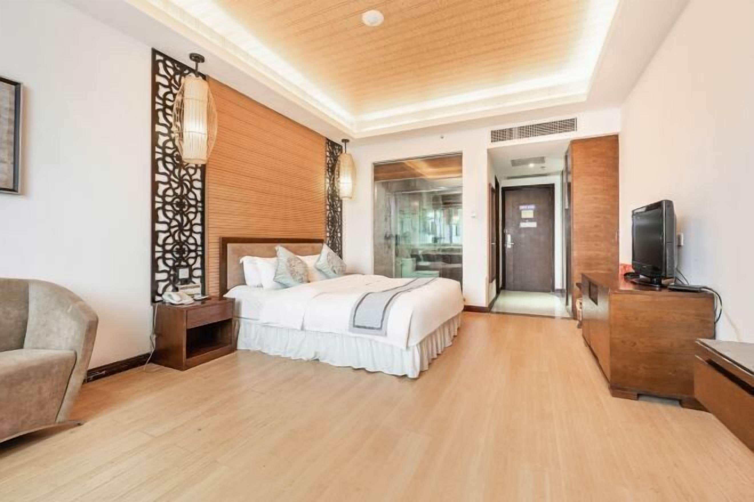 Una habitación de hotel moderna con techo de madera, una cama con ropa blanca, una mesita de noche, un televisor en un mueble de madera y una puerta de vidrio que da a un baño.