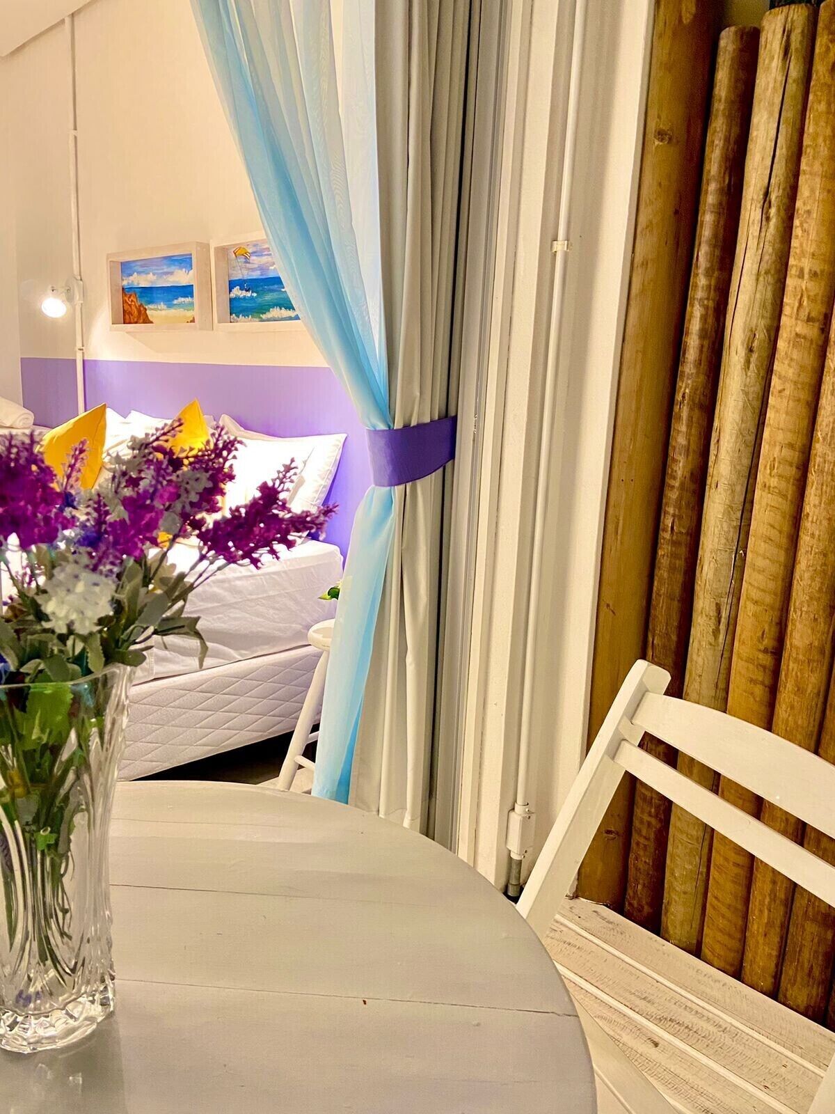 Una habitación con una cama, una mesa con un jarrón de flores y vista a la playa a través de cortinas.
