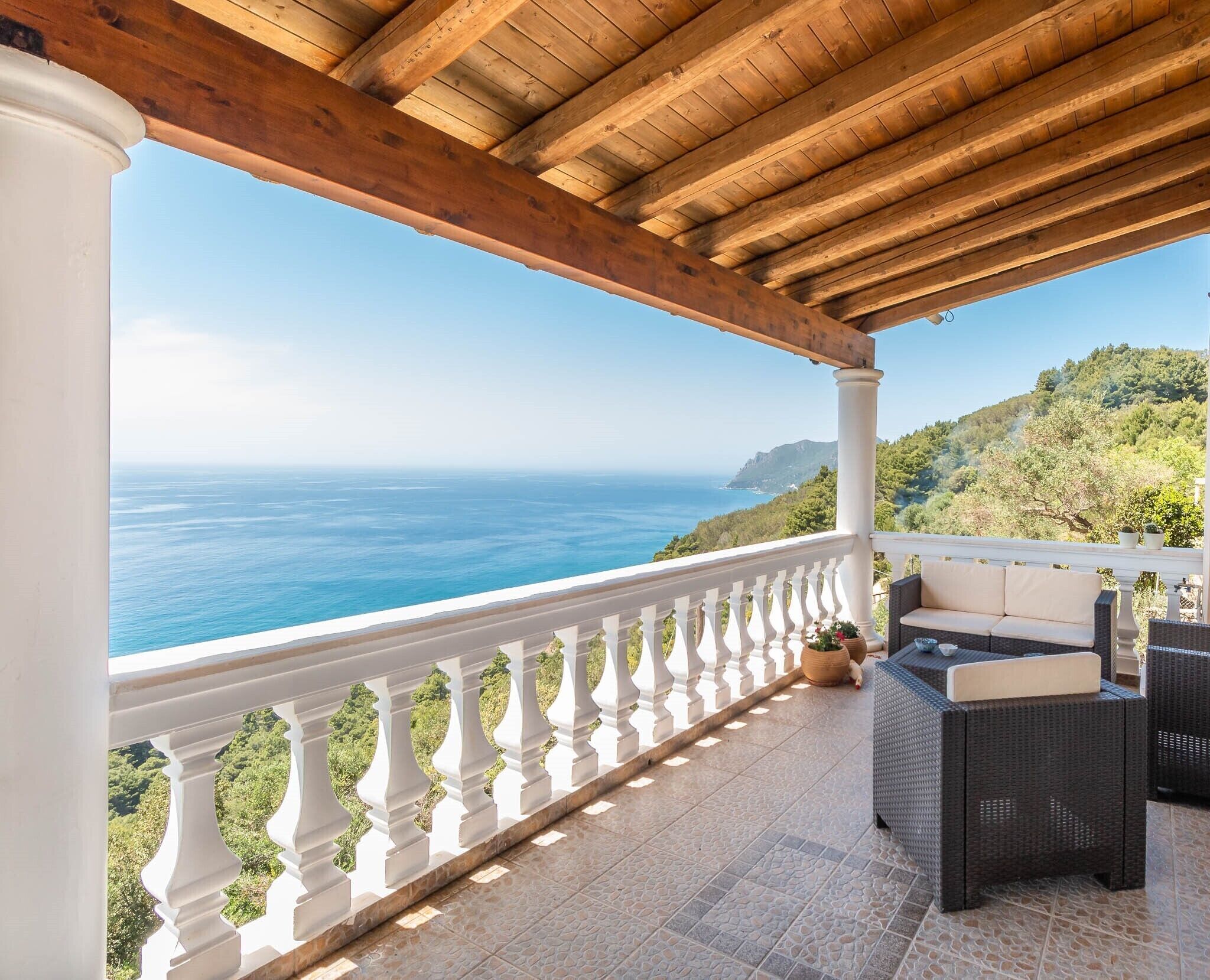 Ein überdachter Außenbereich mit weißer Balustrade, Korbmöbeln und Blick auf Meer und Hügel.