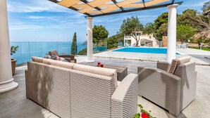 Un área de estar al aire libre con techo, mobiliario de mimbre, piscina y vista al mar.