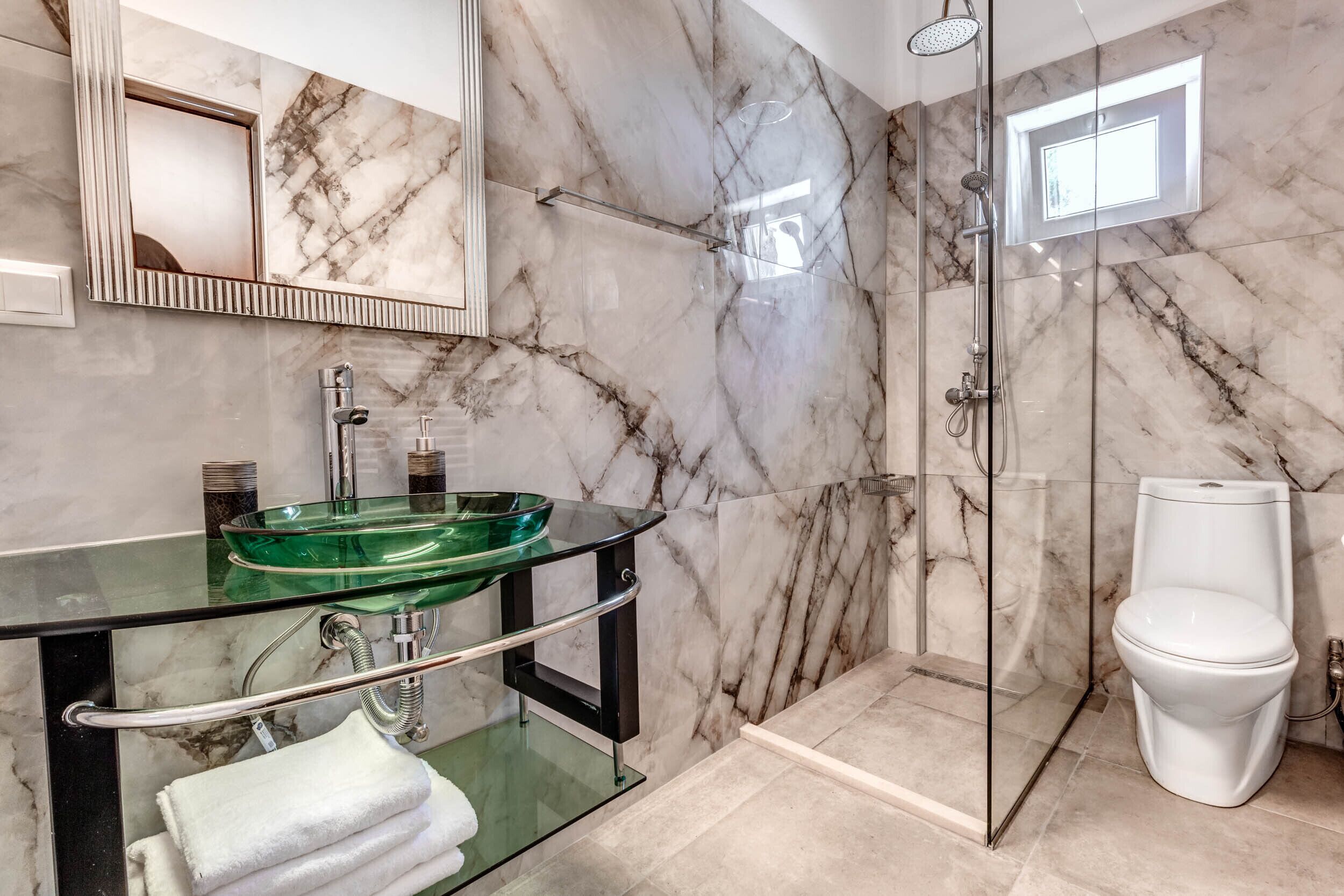 Un bagno moderno con lavabo in vetro, pareti in marmo, doccia con vetro e water bianco.
