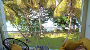 A balcony with a yellow sofa and two chairs, overlooking a grassy area with trees and a view of the sea. - Large, cosy studio with sea view, pool and beach (Le Gosier)