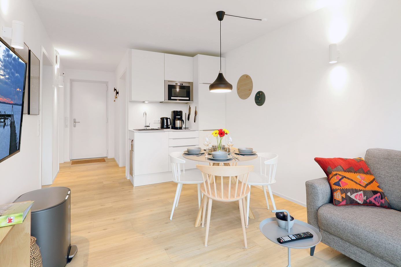 A modern kitchen with white cabinets, a dining table with chairs, and a grey sofa with a colorful pillow.
