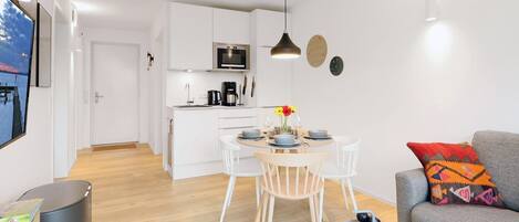 A modern kitchen with white cabinets, a dining table with chairs, and a grey sofa with a colorful pillow.