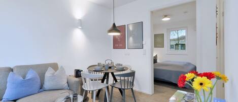 Una sala de estar moderna con sofá, mesa de comedor, sillas, una mesita auxiliar con control remoto y un jarrón con flores.
