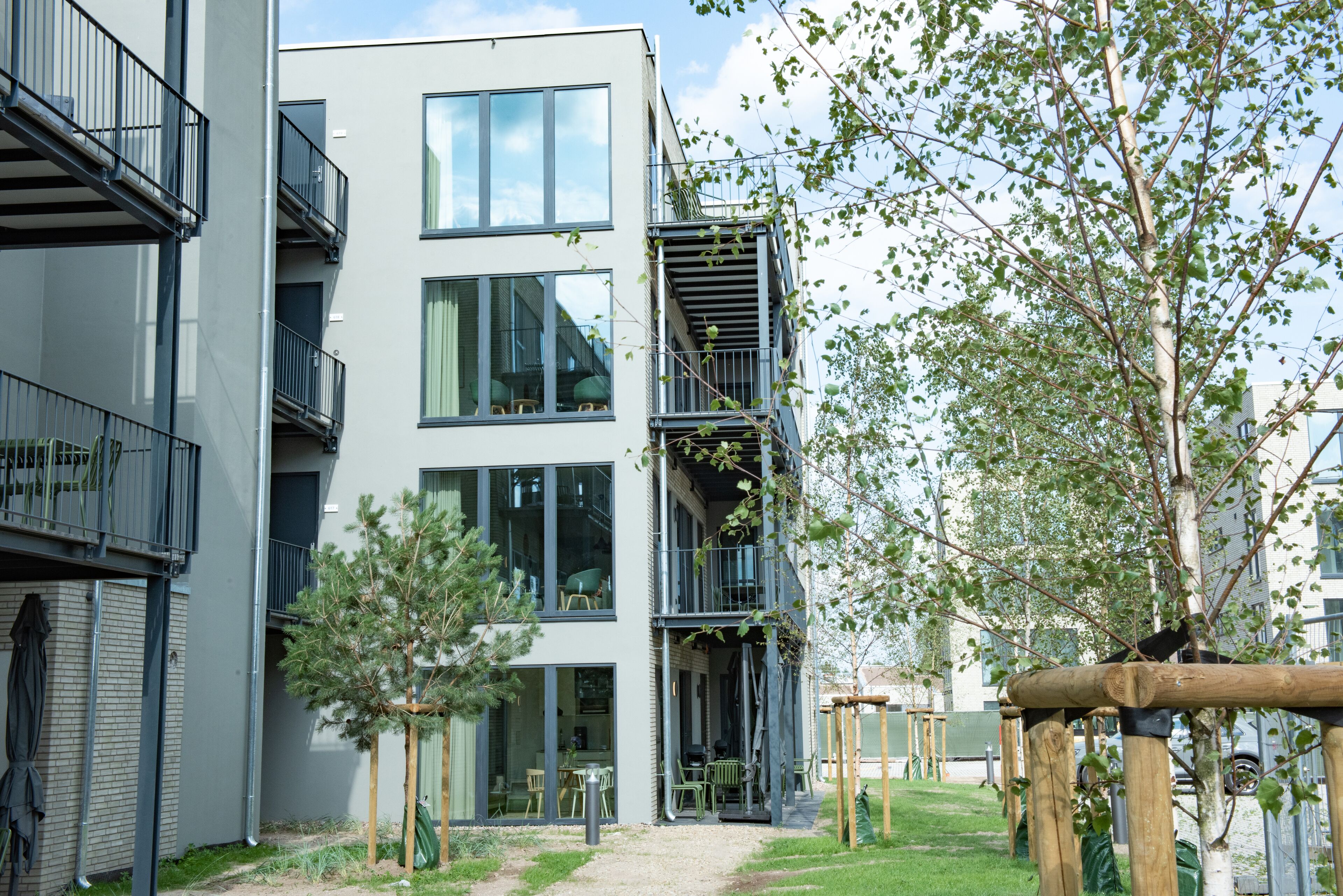 A modern multi-story residential building with balconies, a small garden area with trees, and outdoor seating.