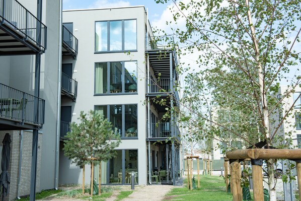 A modern multi-story residential building with balconies, a small garden area with trees, and outdoor seating. - Marissa Resort (Lembruch)