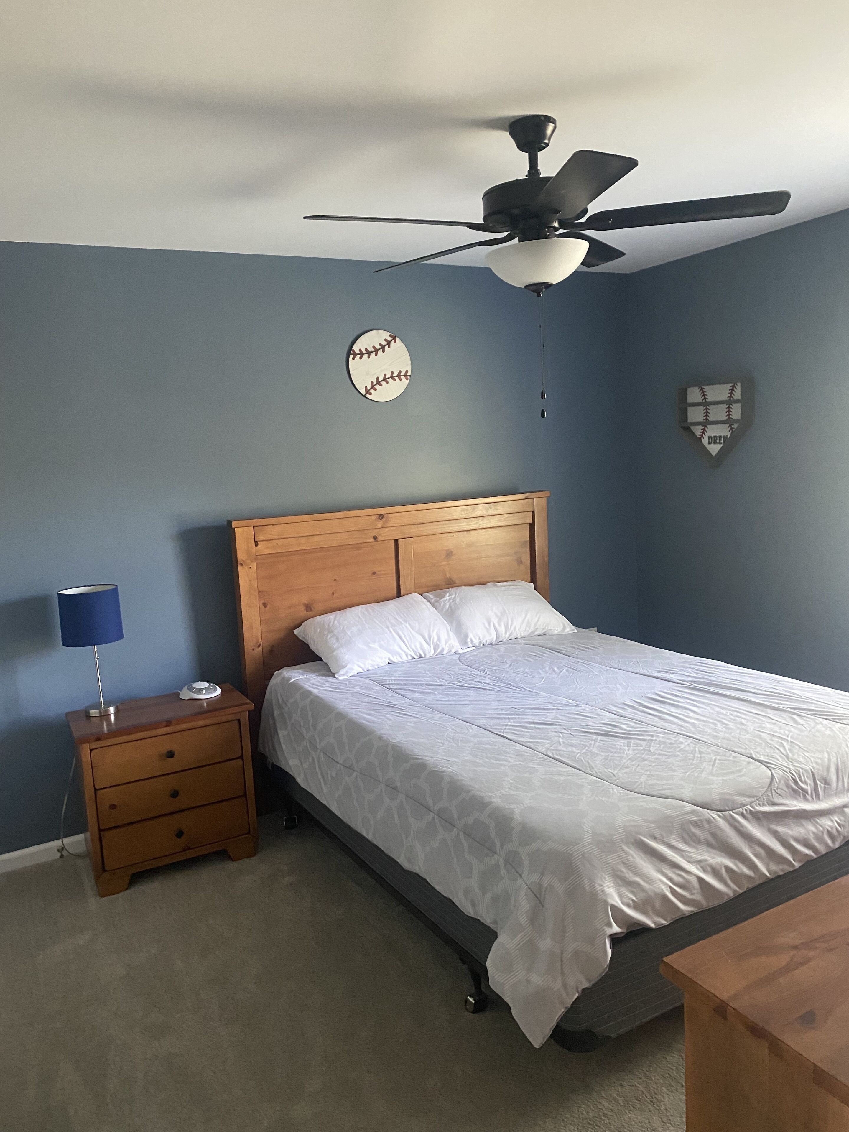 Quarto com cama, mesa de cabeceira, luminária, ventilador de teto e decoração na parede.