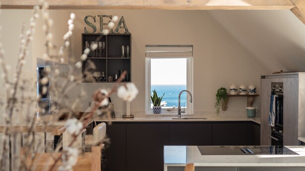A modern kitchen with a view of the sea, featuring dark cabinetry, a window with a view, and a decorative sign above the sink. - Church Rooms (Port Isaac)