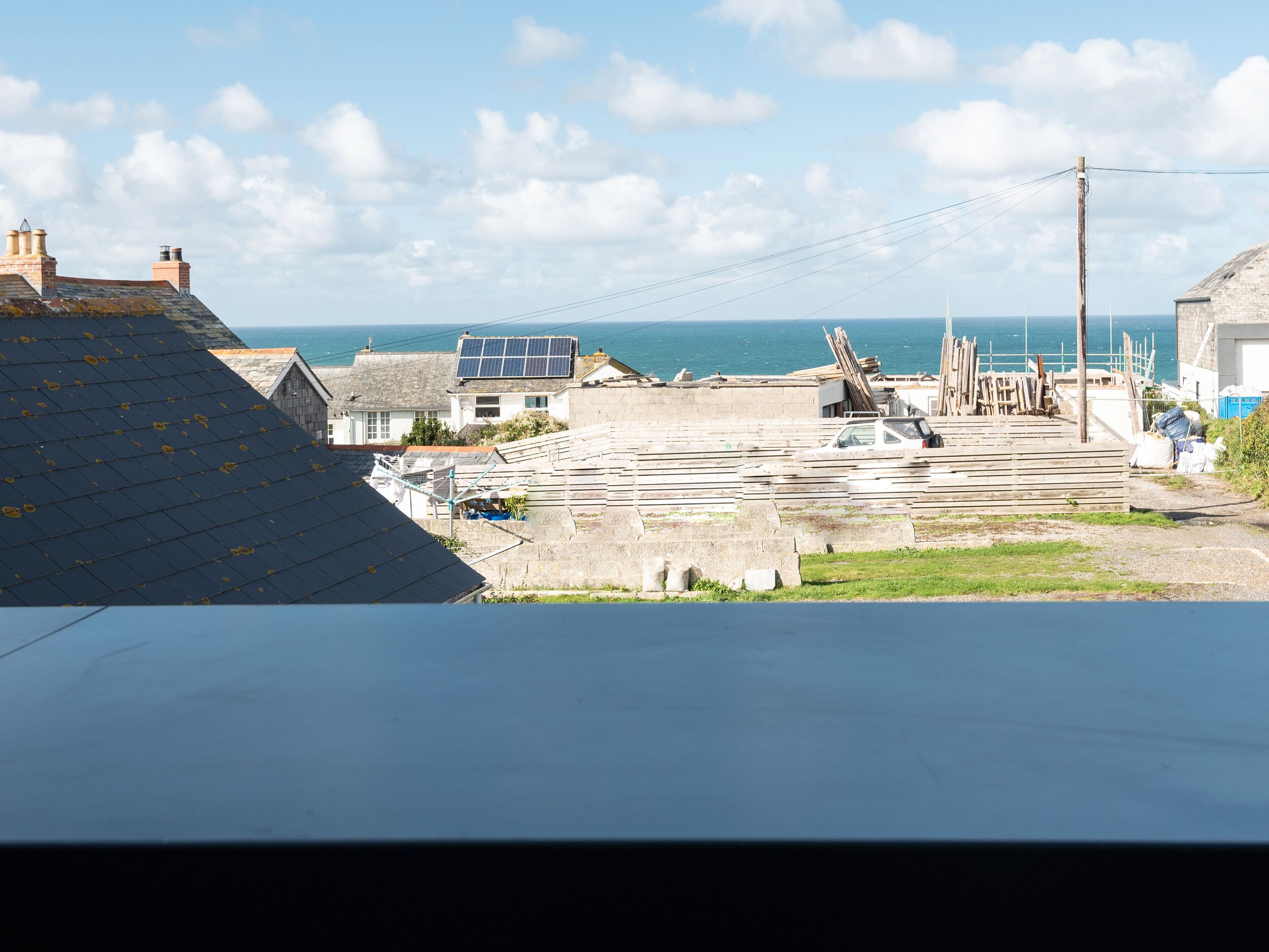 A coastal town with a clear view of the sea, a parking area with stacked wooden planks, and a building with solar panels.
