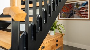 A staircase with wooden steps and black metal railings, a wooden side table with a plant, and a colorful abstract painting on the wall. - Church Rooms (Port Isaac)
