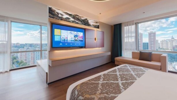 Ein modernes Hotelzimmer mit einem großen an der Wand montierten Fernseher, einer Couch, einem Bett mit gemustertem Überwurf und großen Fenstern mit Blick auf die Stadt.