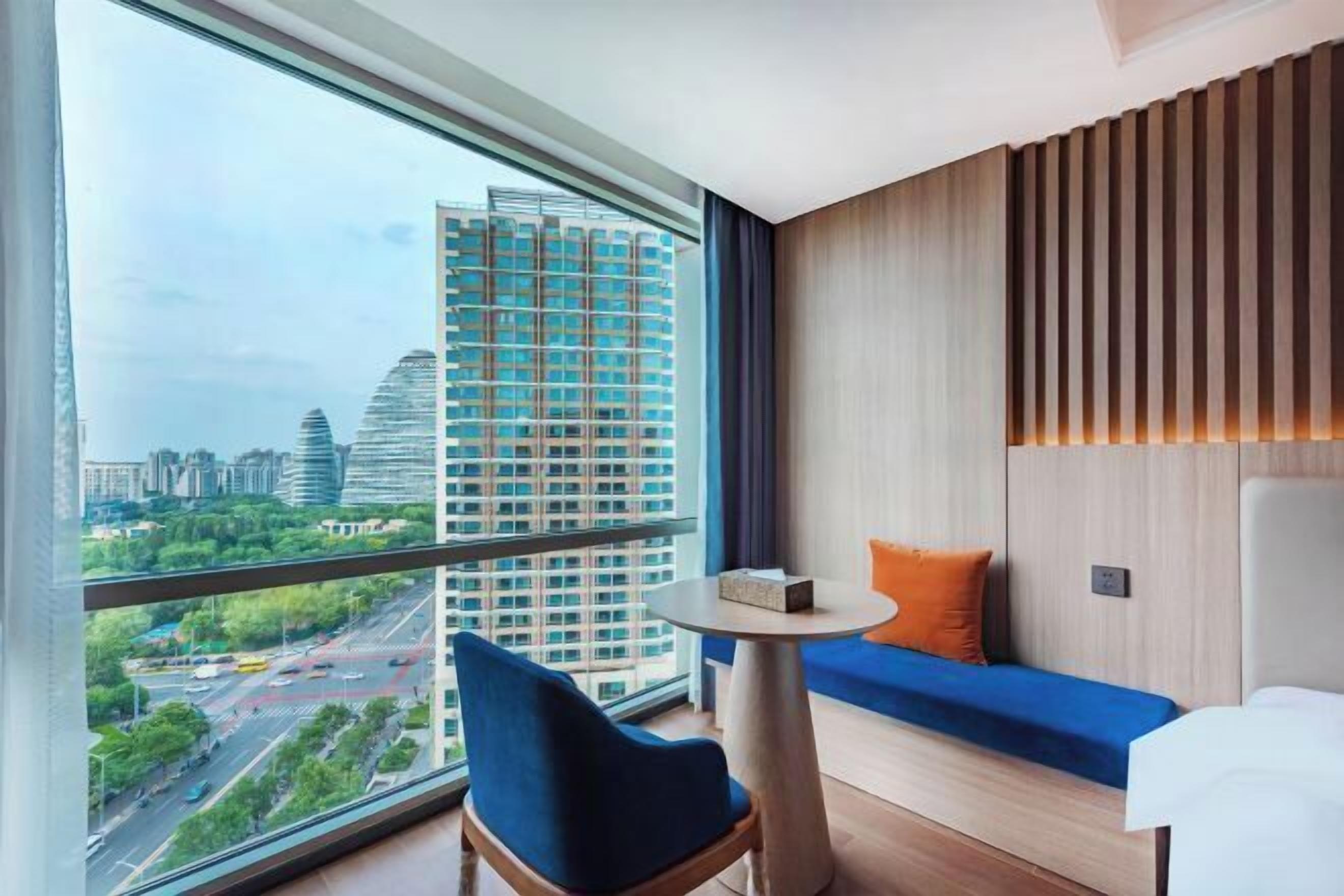Habitación de hotel moderna con un gran ventanal que ofrece vistas a la ciudad, una pequeña mesa redonda, un sofá con un cojín azul y una pared de acento de madera.