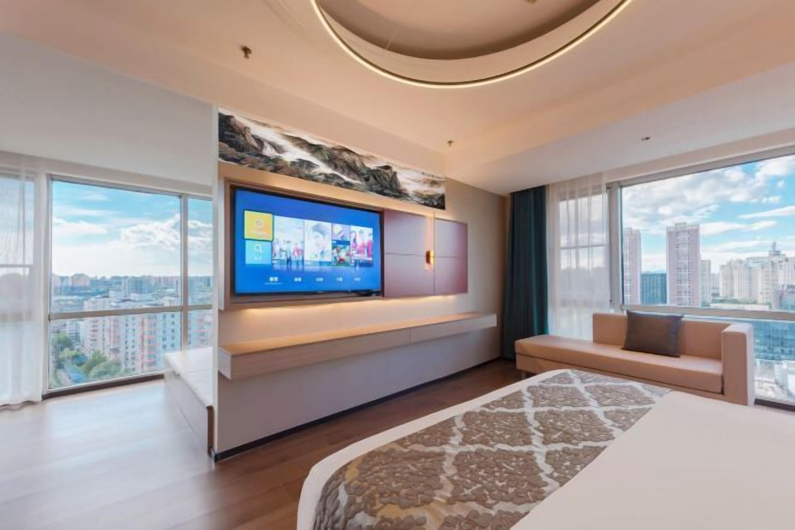 A modern hotel room with a large TV mounted on the wall, a sofa, a bed with a patterned bedspread, and large windows offering a city view.