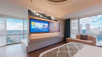 A modern hotel room with a large TV mounted on the wall, a sofa, a bed with a patterned bedspread, and large windows offering a city view.