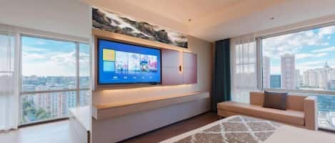Une chambre d’hôtel moderne dotée d’un grand téléviseur fixé au mur, d’un canapé, d’un lit avec une couvre-lit à motifs et de grandes fenêtres offrant une vue sur la ville.