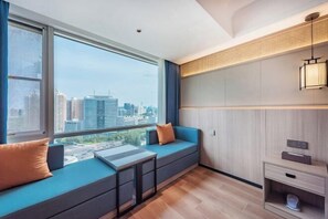 Ett modernt hotellrum med ett stort fönster som erbjuder utsikt över staden, en inbyggd bänk med förvaringsutrymme och ett litet nattduksbord.
