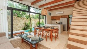 A modern kitchen with wooden beams, a dining area with a table set for four, and a large window overlooking a stone wall and greenery. - Aldea Zama Jungle Home: Private Pool & Garden Tub (Tulum)