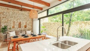 A modern kitchen with a stone wall, large windows, and a dining area with a wooden table and chairs. - Aldea Zama Jungle Home: Private Pool & Garden Tub (Tulum)