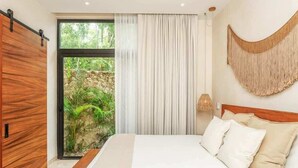 A bedroom with a bed, a sliding door, a view of greenery, and a wall-mounted light fixture. - Aldea Zama Jungle Home: Private Pool & Garden Tub (Tulum)