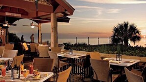 An outdoor dining area with tables and chairs, overlooking a body of water and palm trees during sunset. - 562 Beachy Keen Cottage| New Pool | Mins 2 Beaches (Naples)
