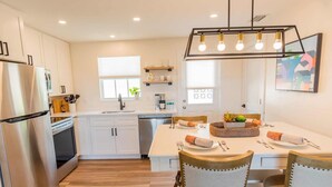 A modern kitchen with white cabinets, a stainless steel refrigerator, a built-in dishwasher, and a dining area with a wooden table and chairs. - 562 Beachy Keen Cottage| New Pool | Mins 2 Beaches (Naples)