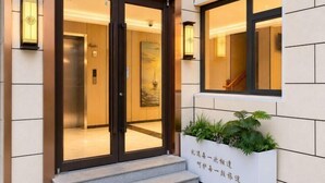 Entrada de hotel con puerta de cristal, una escalera y una jardinera con follaje.