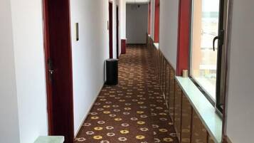 Un couloir d’hôtel avec un tapis à motifs, des portes rouges et des fenêtres donnant sur un paysage agréable.