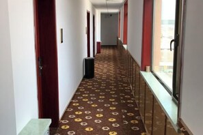 Un couloir d’hôtel avec un tapis à motifs, des portes rouges et des fenêtres donnant sur un paysage agréable.