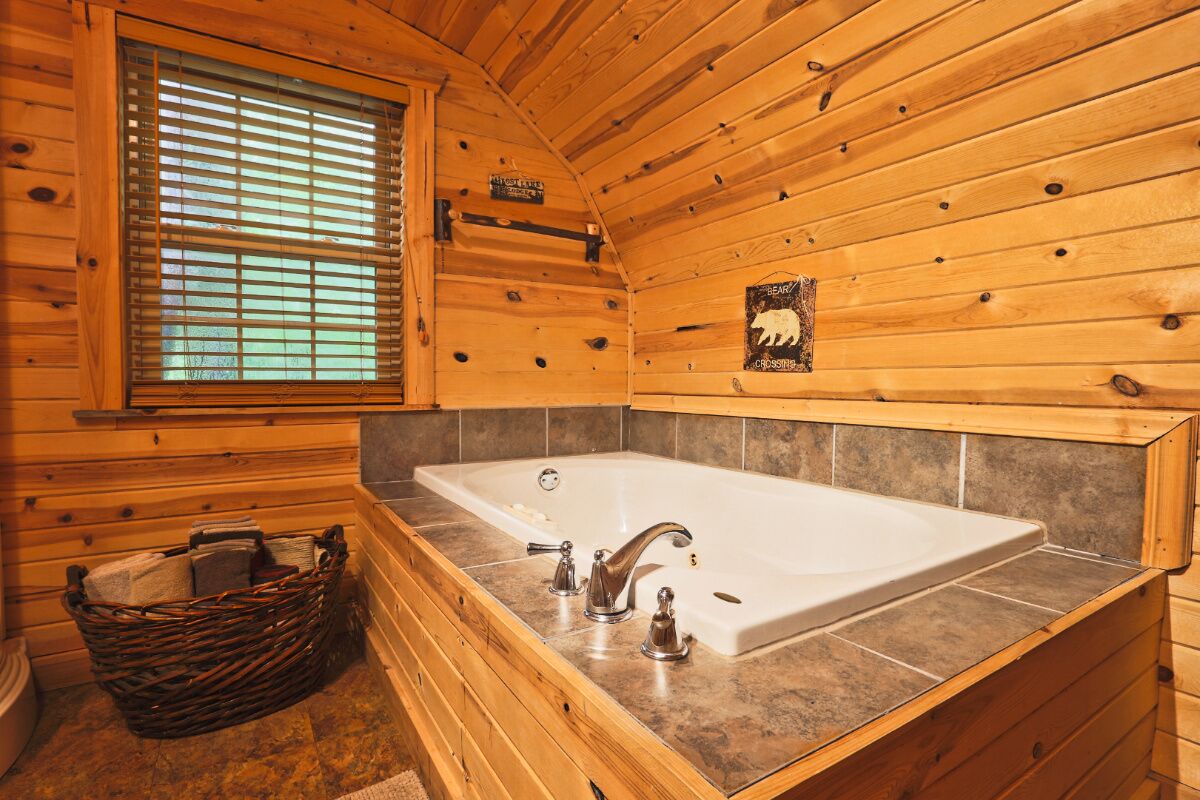A wooden cabin bathroom with a large bathtub, a window with blinds, and a bear-themed wall decoration.