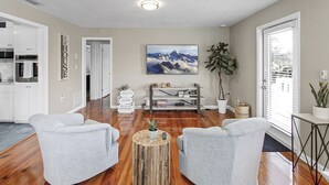 A modern living room with a wooden floor, grey sofas, a coffee table with a plant, a TV on the wall, and a kitchen in the background. - Delightful 2/2 Beach House LBK 5 Min drive to Beach (Longboat Key)