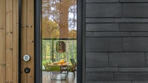 Un interior moderno con una puerta de madera que da a un comedor con mesa y sillas, y vista a un bosque a través de amplios ventanales.