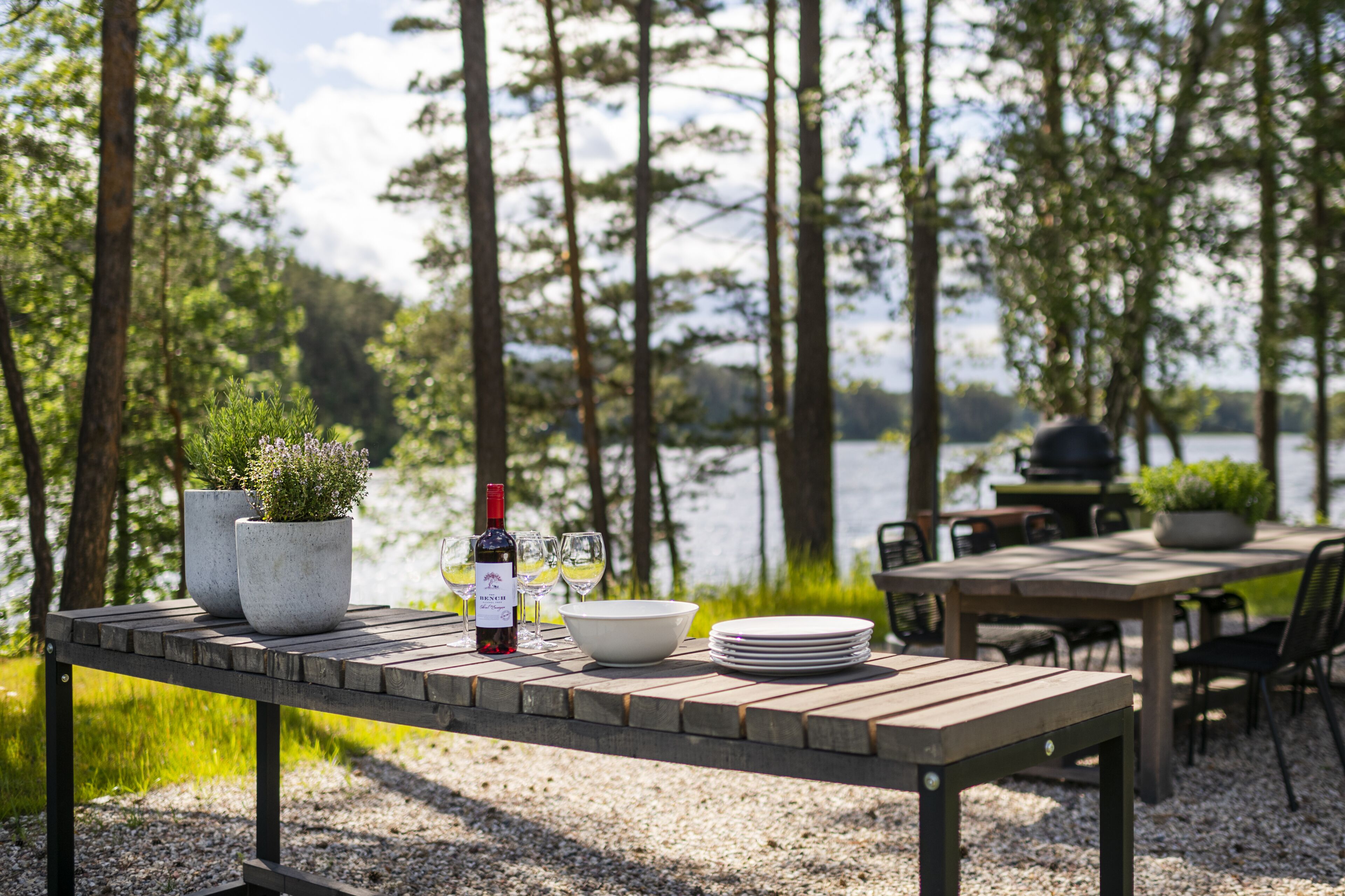 Een houten tafel gedekt met een wijnfles, glazen en een schaal, omringd door stoelen en een barbecue, met een meer en bomen op de achtergrond.