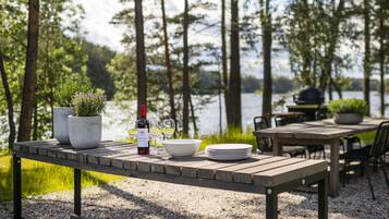 Een houten tafel gedekt met een wijnfles, glazen en een schaal, omringd door stoelen en een barbecue, met een meer en bomen op de achtergrond.