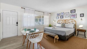 A bedroom with a bed, a bedside table, a lamp, a window with curtains, a small table with chairs, and a wall with framed pictures. - Perfect Location Vinoy Beach Getaway Sun and fun (St. Petersburg)