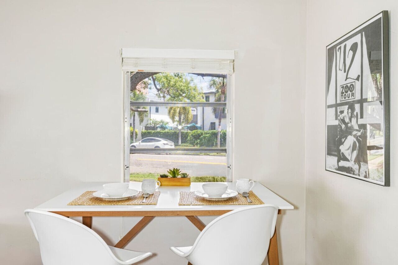 A dining area with a table set for two, a window with a view of a street and buildings, and a framed picture on the wall.