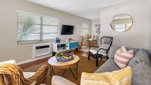 A living room with a grey sofa, wicker chairs, a round coffee table, a TV on the wall, and a large window with blinds. - Vinoy Beach getaway, Fun in the Sun Close to bars! (St. Petersburg)