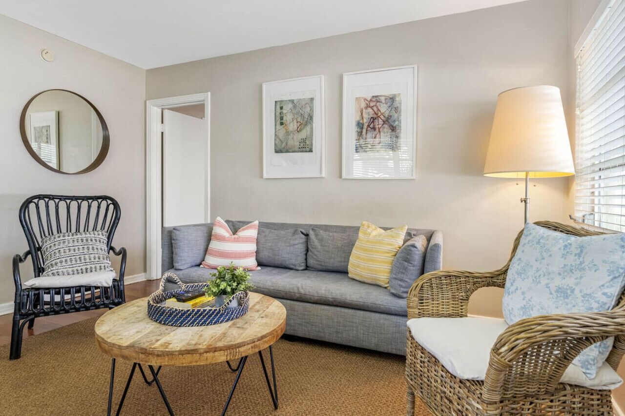 A living room with a grey sofa, a wicker chair, a round wooden coffee table, and a floor lamp. There are two framed artworks on the wall and a mirror above the sofa.