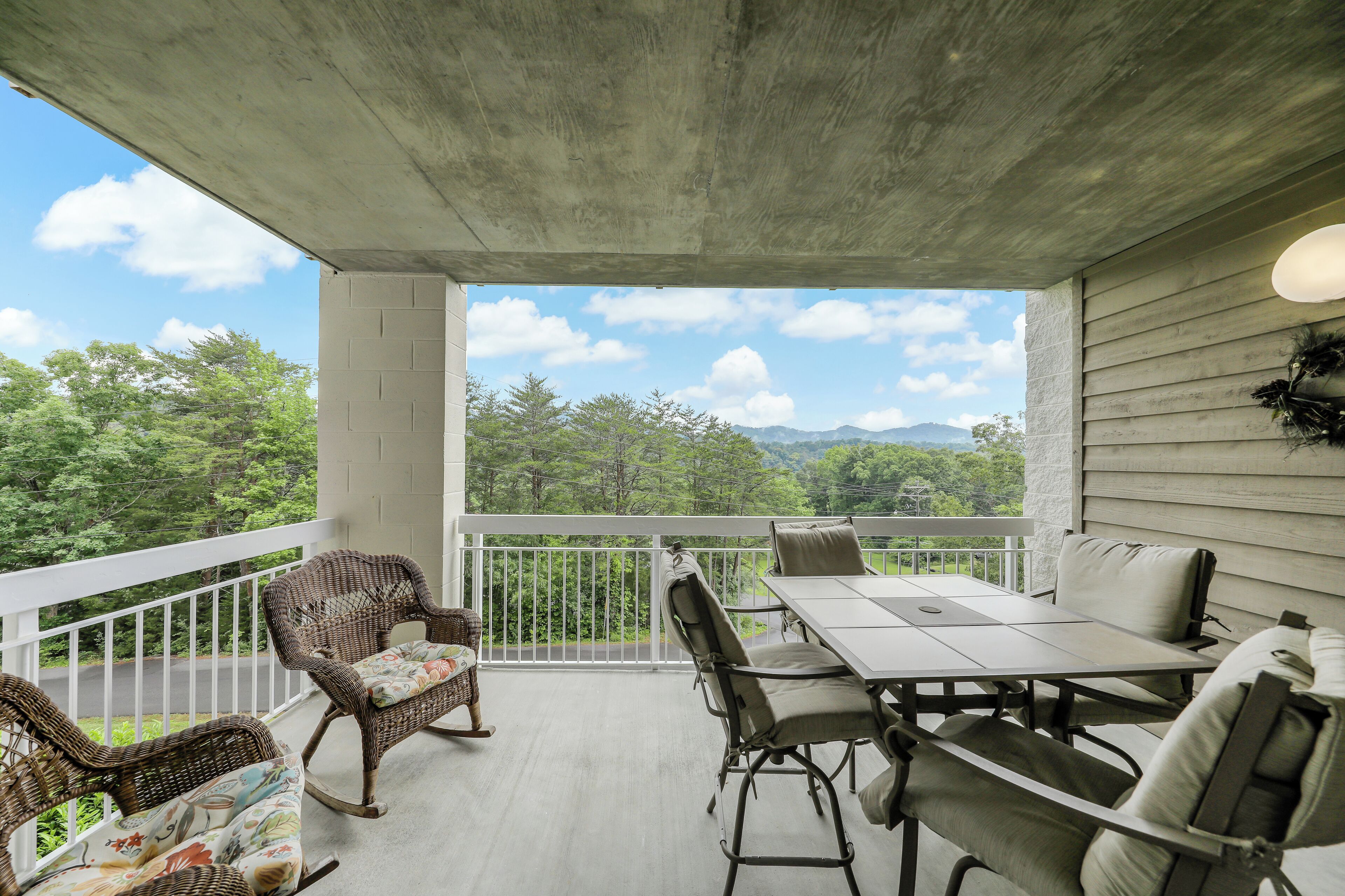 A covered outdoor patio with wicker furniture and a table, overlooking a scenic view of trees and mountains.