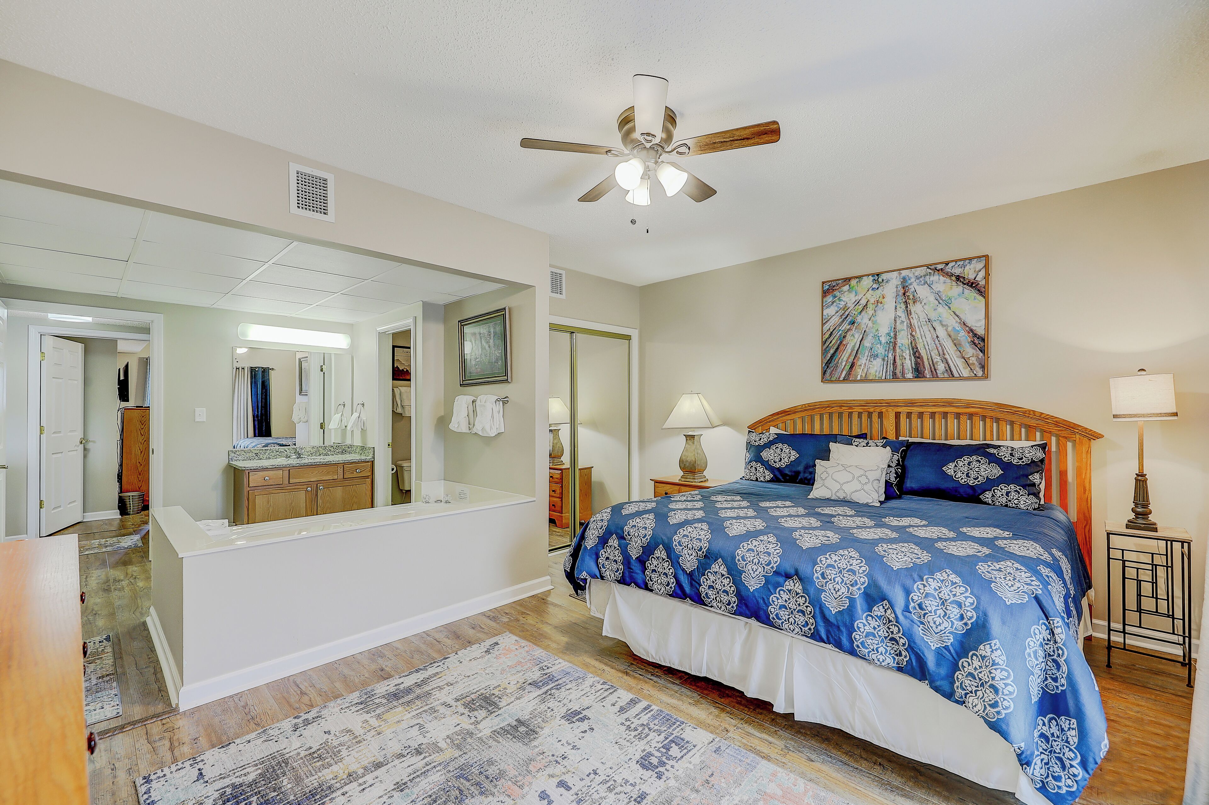 A bedroom with a large bed, a ceiling fan, a bedside table with a lamp, a mirror, and a bathroom visible through an open door.