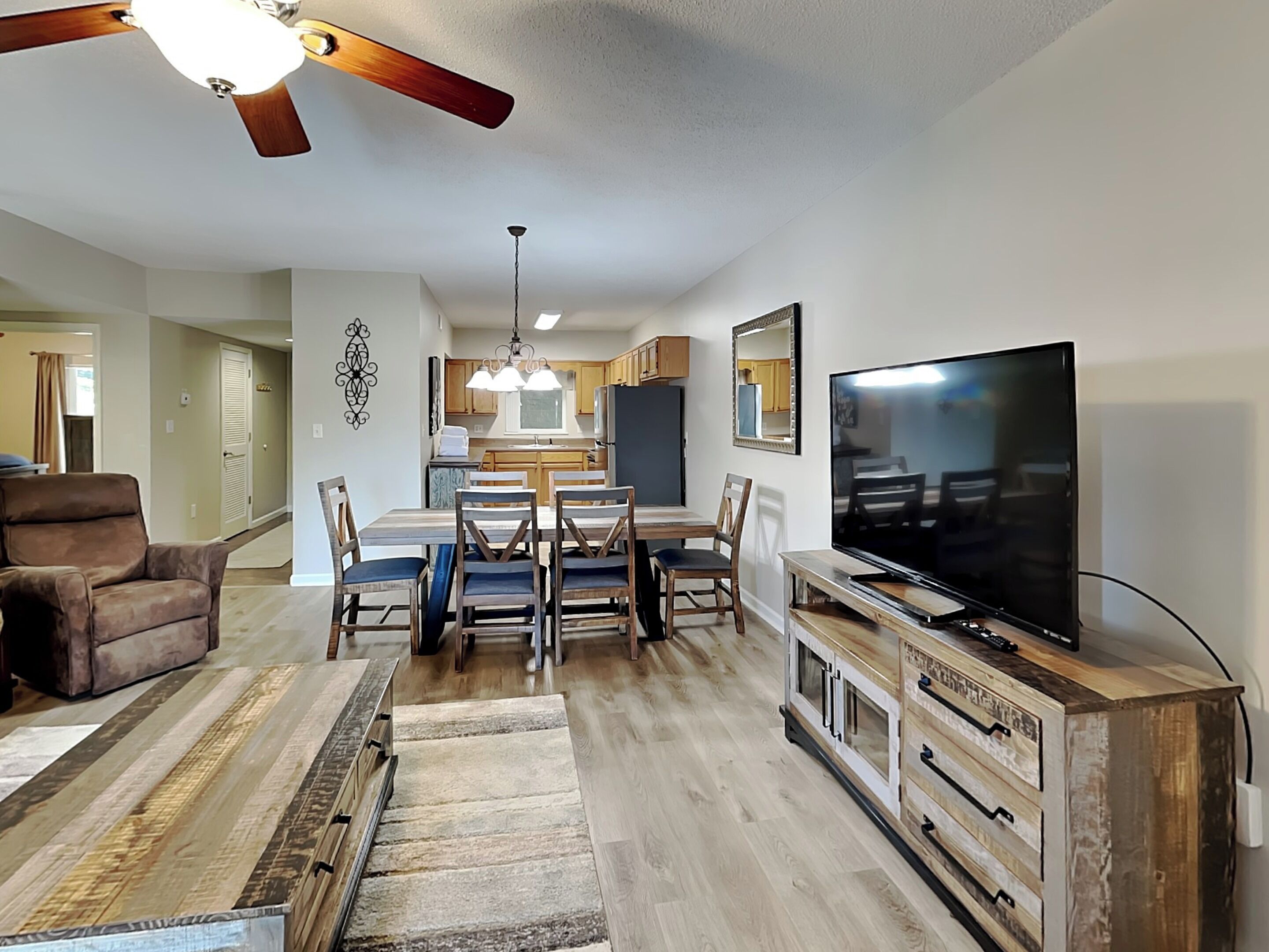 A modern living room with a wooden dining table, comfortable seating, a flat-screen TV, and a ceiling fan.