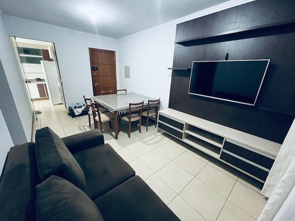 A modern living room with a dark-colored sofa, a dining table with chairs, and a flat-screen TV mounted on a dark wall.