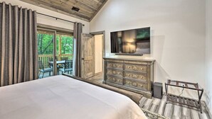 A bedroom with a large bed, a dresser, a TV mounted on the wall, and a view of the outdoors.