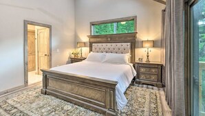 A bedroom with a large bed, wooden headboard, two bedside tables with lamps, a window with a view of greenery, and a door leading to another room.