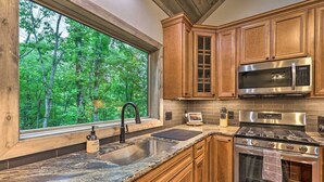 A kitchen with wooden cabinets, a stainless steel stove, a microwave, and a window overlooking a forest.